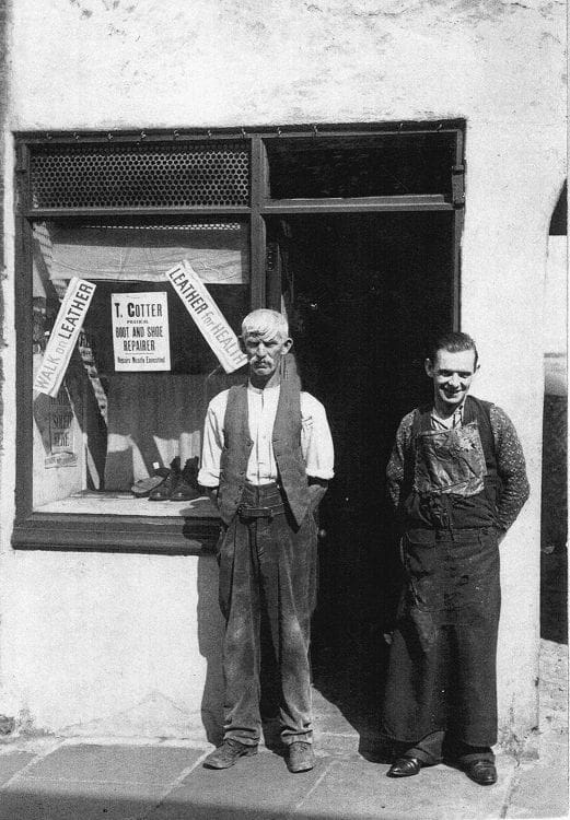 Black and white photograph showing two boot and shoe repairers stood outside their shop