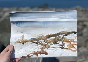 Woman holding watercolour painting of coastal scene in foreground, with coast in background