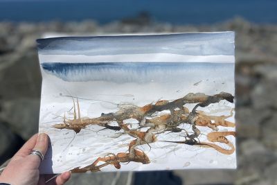 Woman holding watercolour painting of coastal scene in foreground, with coast in background