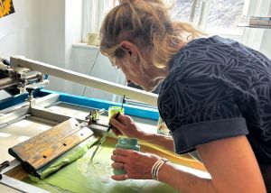 Image shows a woman screen printing with light green ink in a studio space
