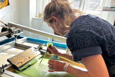 Screen Printing Course Image shows a woman screen printing with light green ink in a studio space